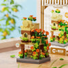 A miniature wooden building set featuring a cabin and windmill, decorated with green foliage and orange flowers.