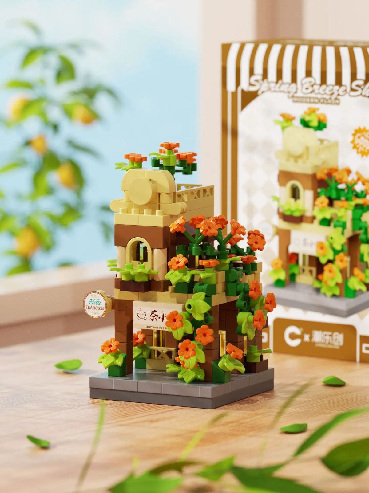A miniature wooden building set featuring a cabin and windmill, decorated with green foliage and orange flowers.