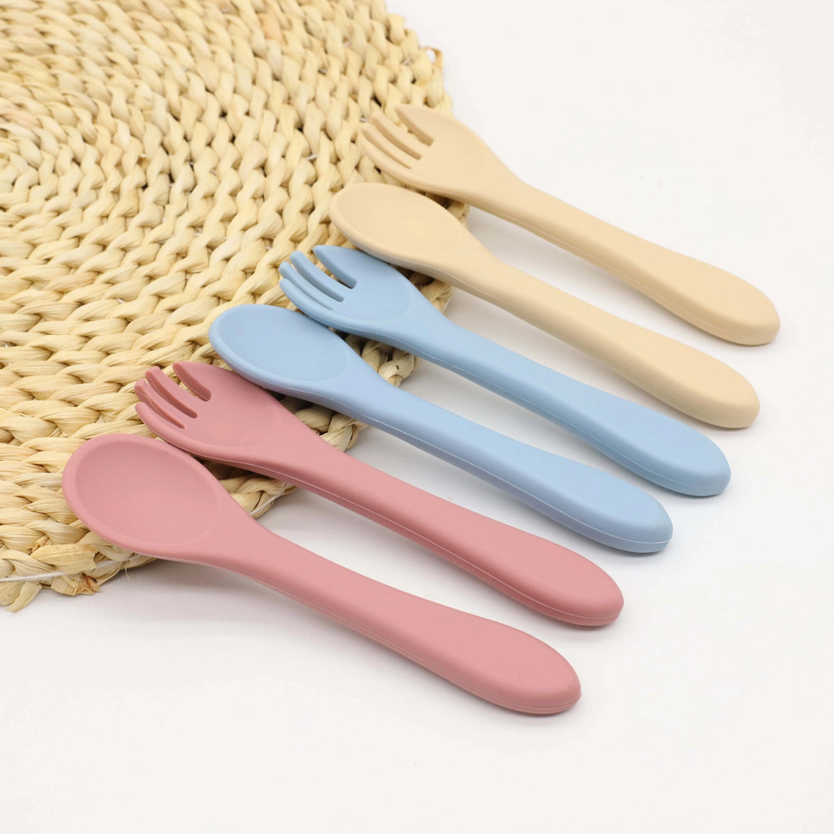 A set of nine silicone baby utensils in various pastel colors, including pink, blue, and beige, resting on a woven mat.