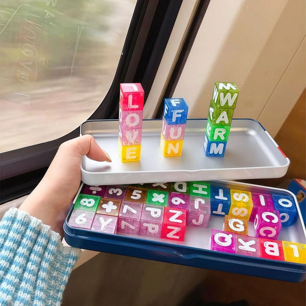 A hand holds a tray of colorful magnetic building blocks for kids, including letters and numbers, next to a window.