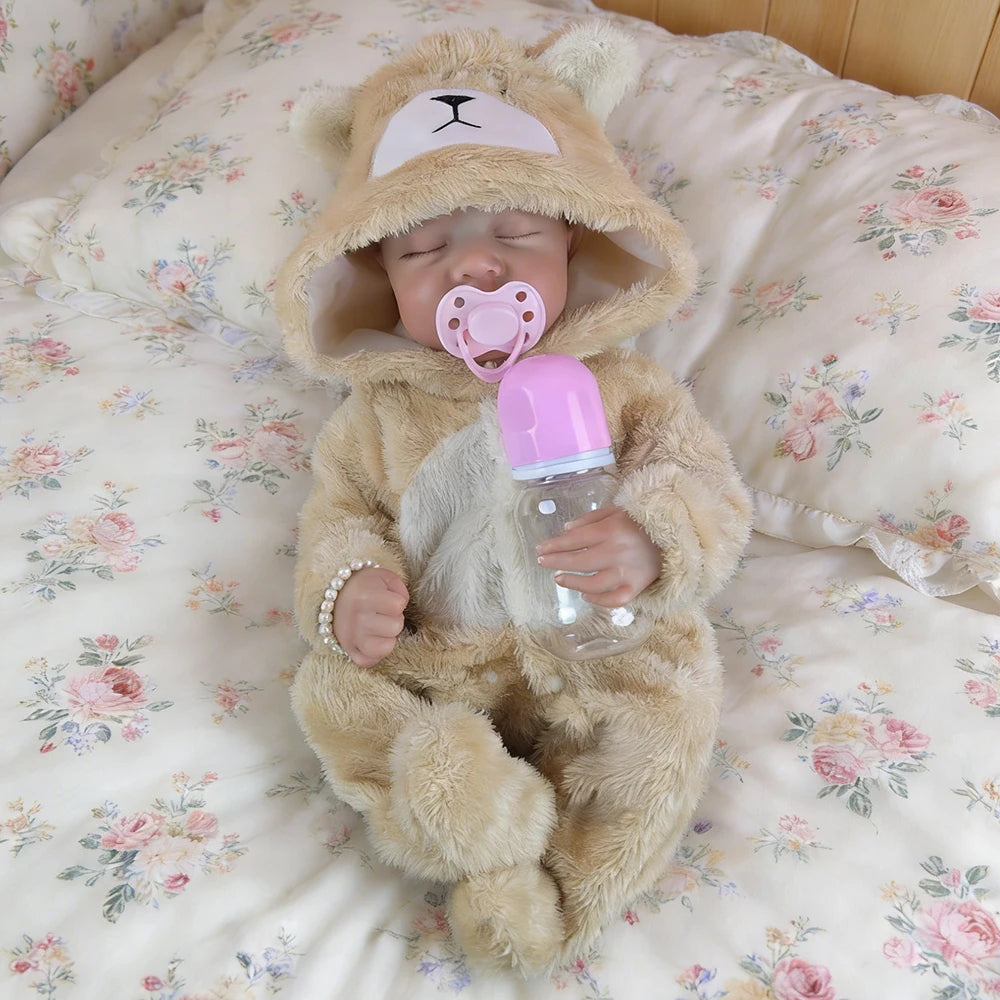 A realistic vinyl reborn baby doll wearing a fluffy bear outfit and holding a pink bottle, lying on a floral patterned bed.