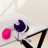 A purple hand crank yarn winder is shown on a white table next to a pink ball of yarn.