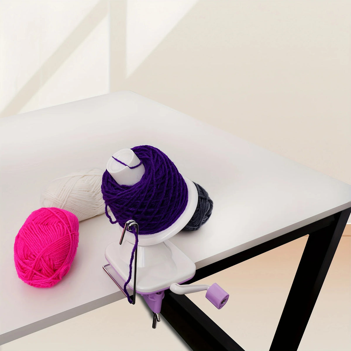 A purple hand crank yarn winder is shown on a white table next to a pink ball of yarn.