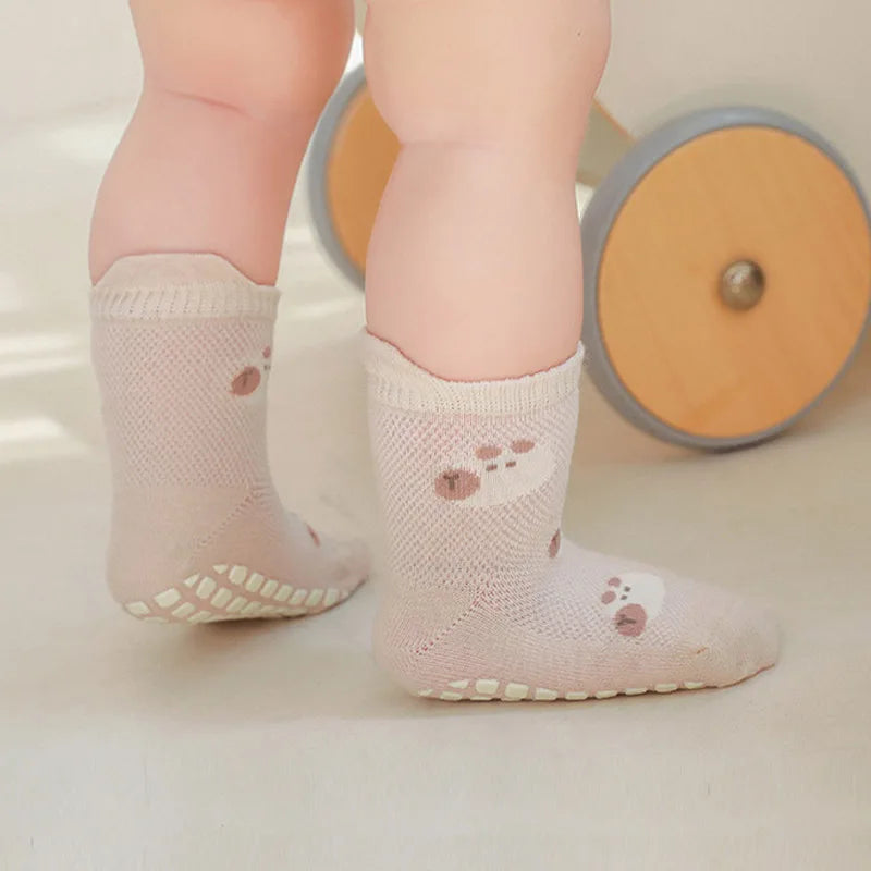 A pair of light pink non-slip cartoon socks for a baby's foot, shown next to a wooden toy.