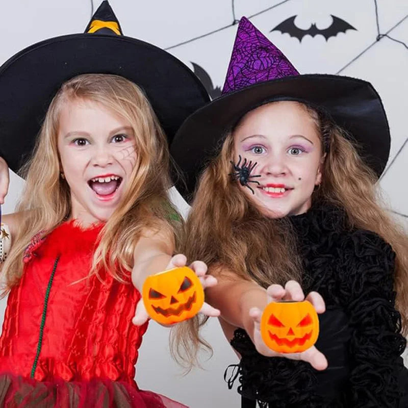Two young girls wearing witch hats holding orange pumpkin and black skeleton squeeze stress balls