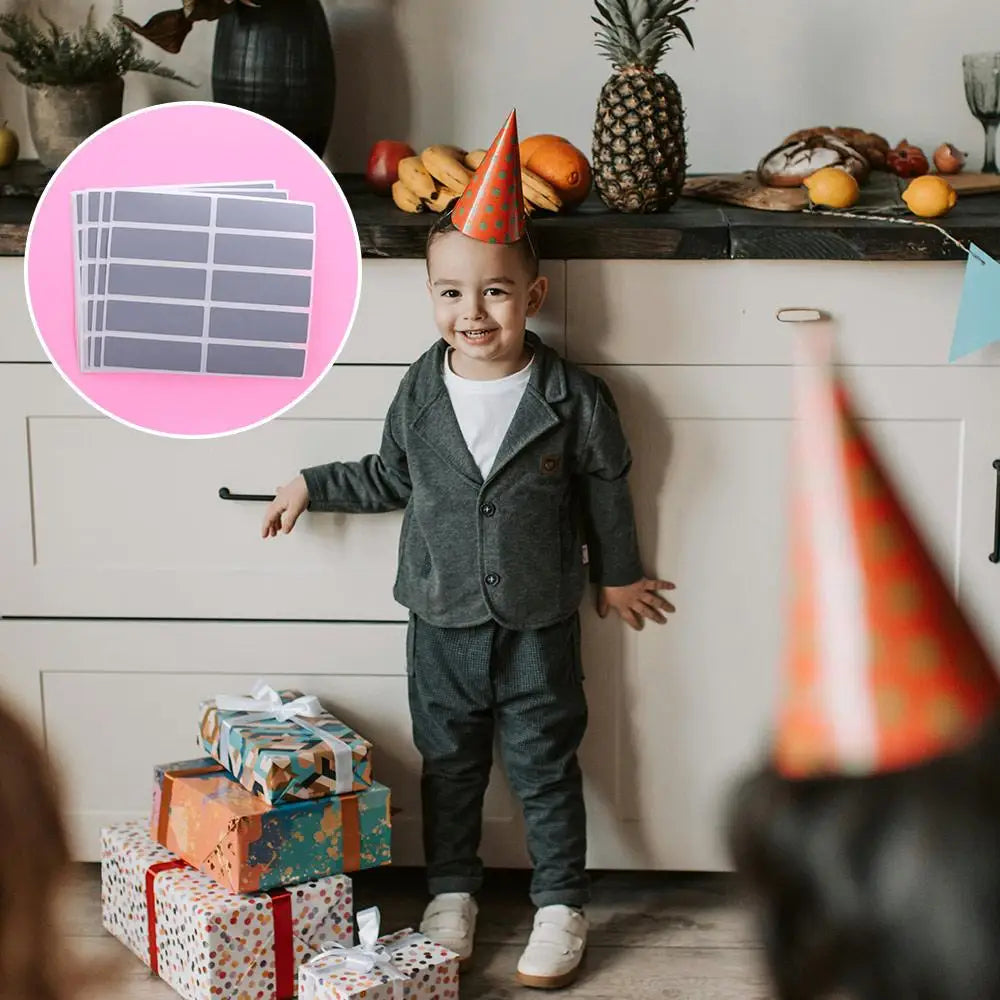 A smiling toddler boy wearing a gray outfit stands in front of a cabinet with various items and colorful gift boxes.