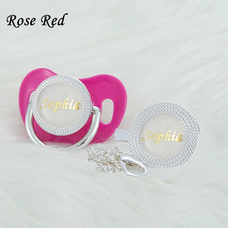A pink silicone pacifier clip set featuring a clear dome with a rhinestone name and a silver clip.