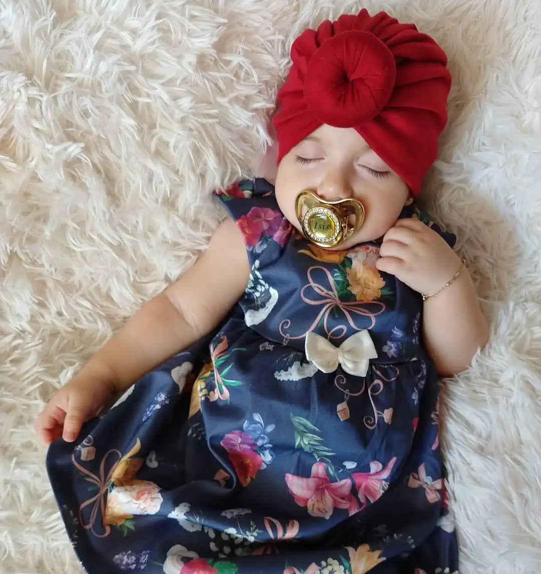 A baby wearing a red turban and a dark floral dress is holding a gold rhinestone pacifier.