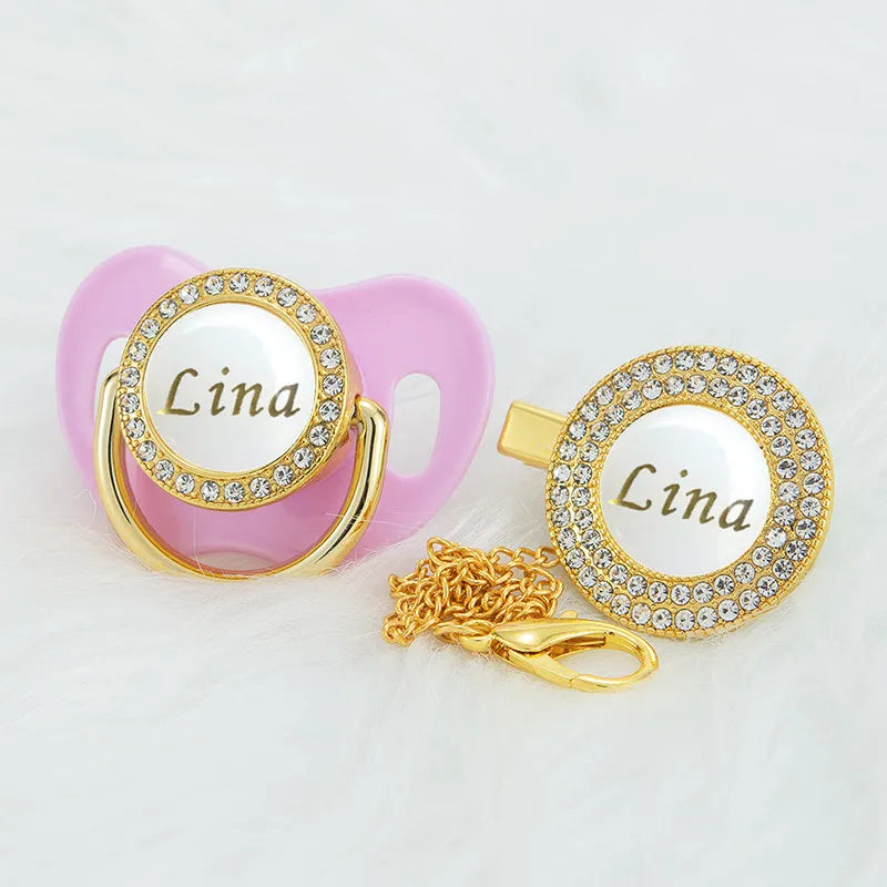 A pink pacifier clip with a gold nameplate and a separate gold chain and pendant.