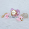 A pink crystal pacifier with a gold clip and a beaded chain featuring the name Lina and a matching decorative charm.