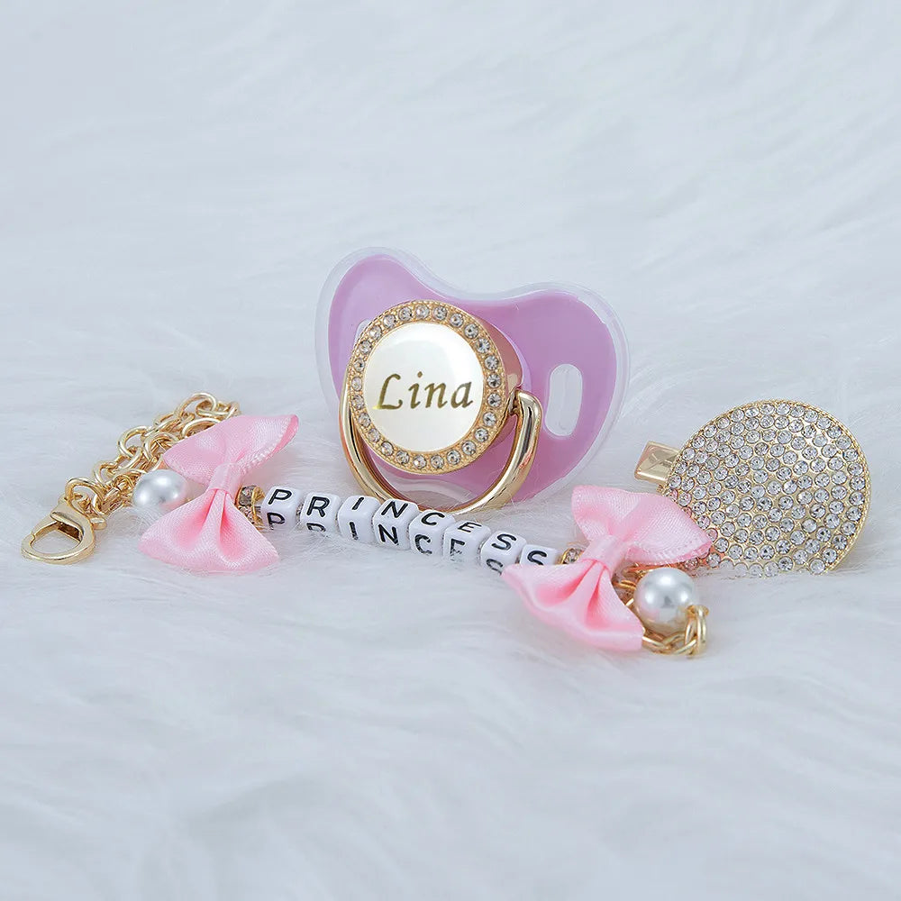 A pink crystal pacifier with a gold clip and a beaded chain featuring the name Lina and a matching decorative charm.