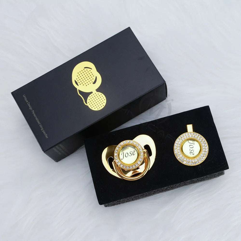 A black box containing a personalized crystal pacifier and clip gift set with gold accents.