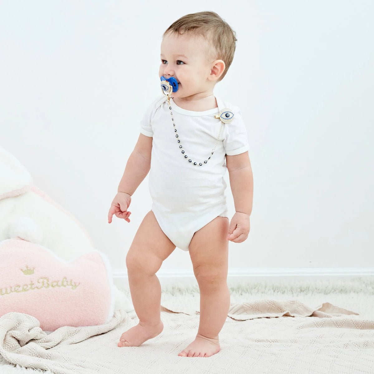 A baby wearing a white outfit with a blue evil eye pacifier and clip set