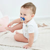 A baby is shown wearing a blue pacifier clip set on their breast and sitting on a light blanket.