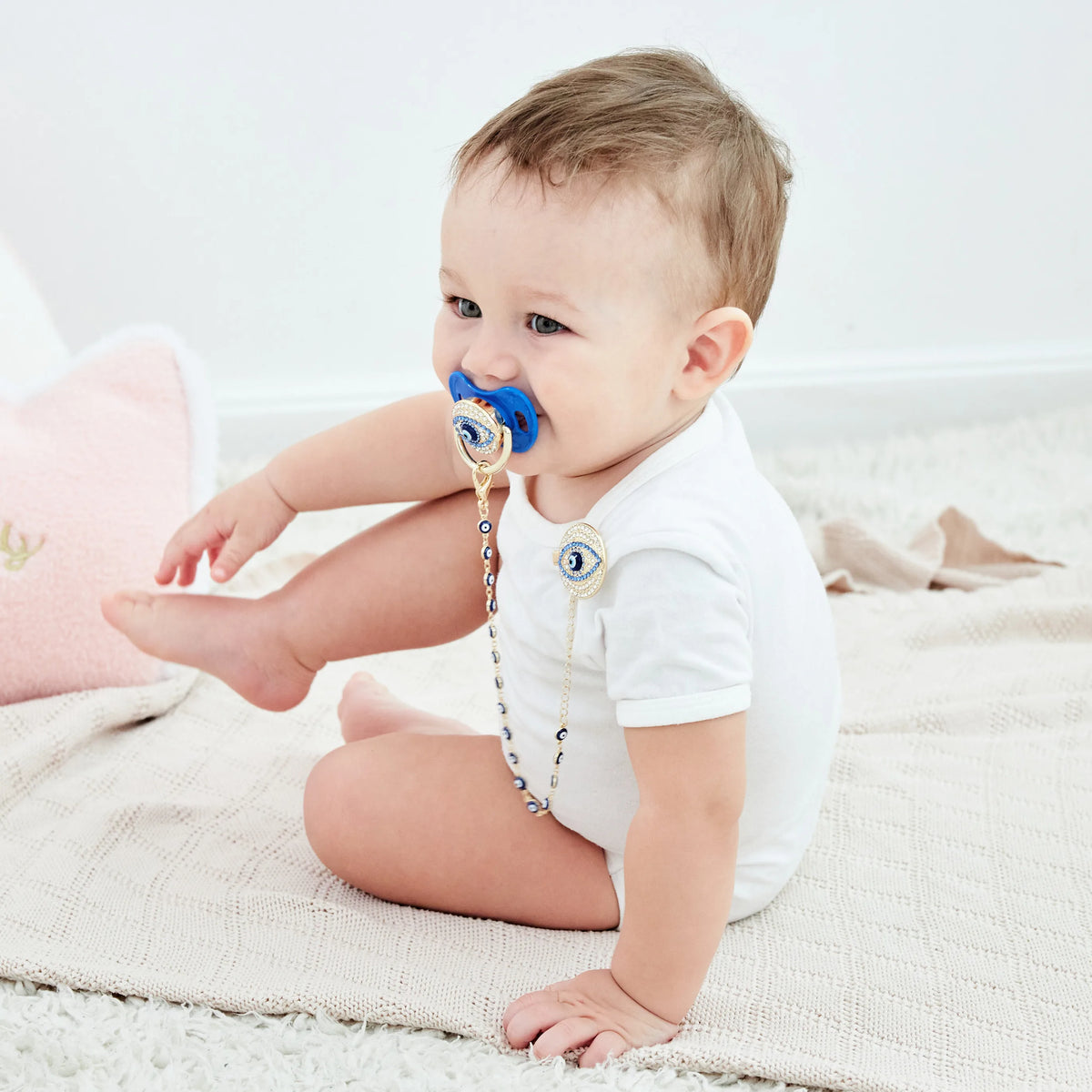 A baby is shown wearing a blue pacifier clip set on their breast and sitting on a light blanket.