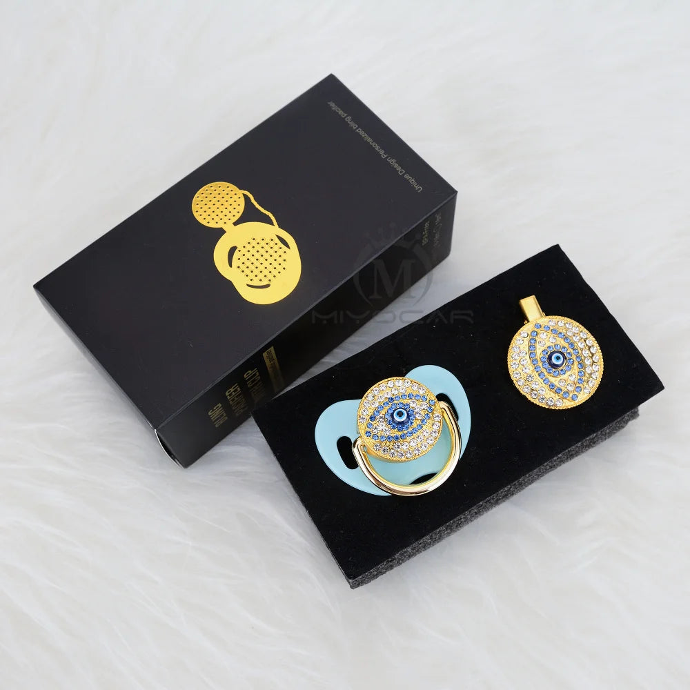 A set of gold-toned evil eye pacifier and clip jewelry displayed in a black box.