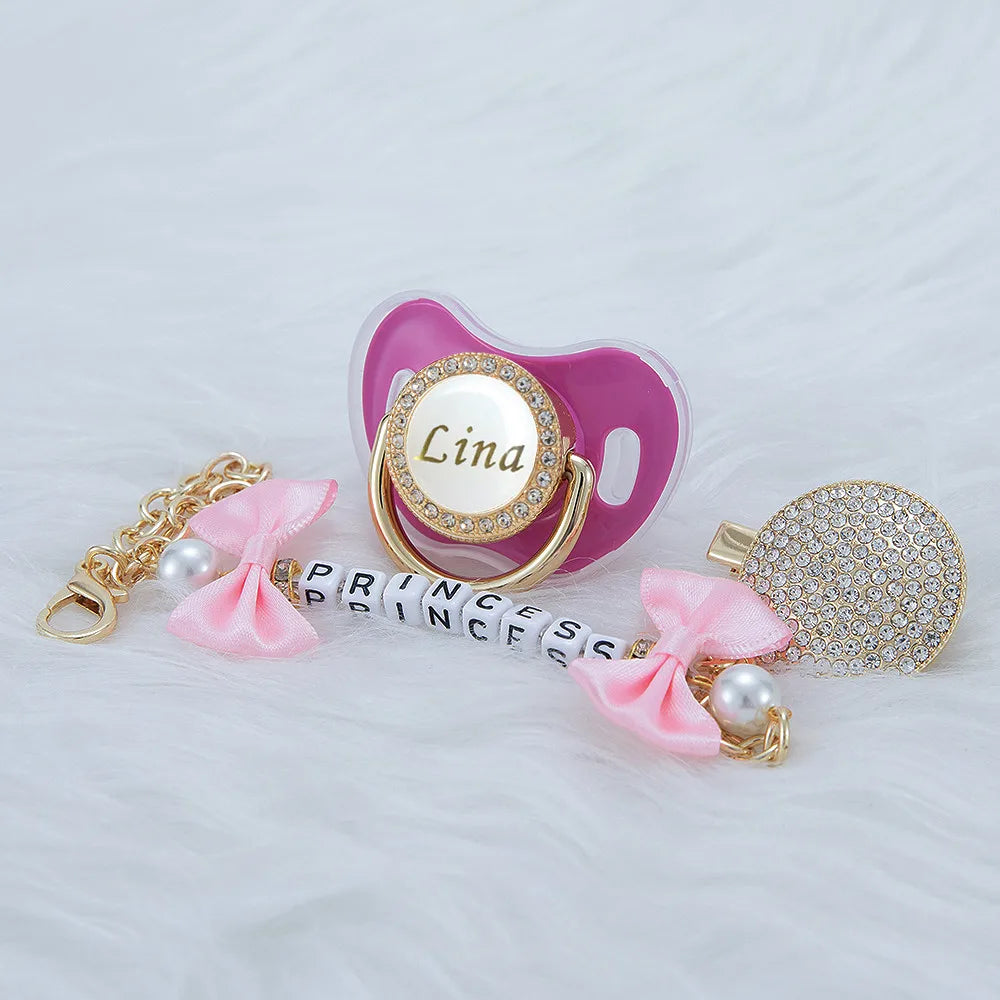 A pink crystal pacifier with a clip and a pearl charm, featuring the name Lina, displayed on white fur.