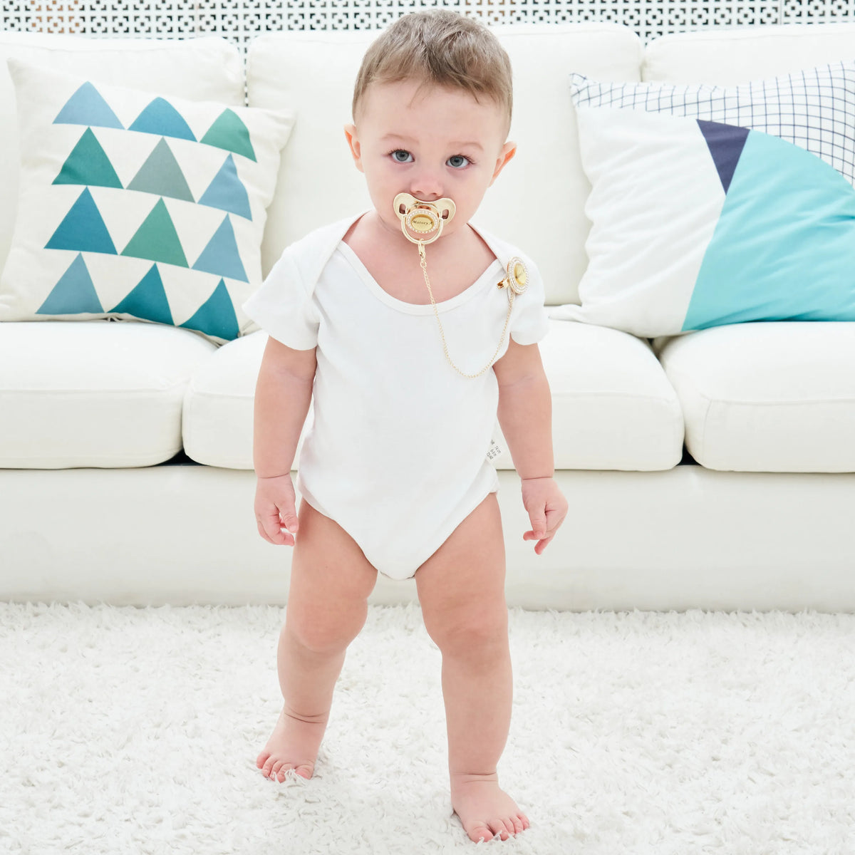 A baby wearing a white onesie with a crystal pacifier and clip on, sitting on a white sofa.
