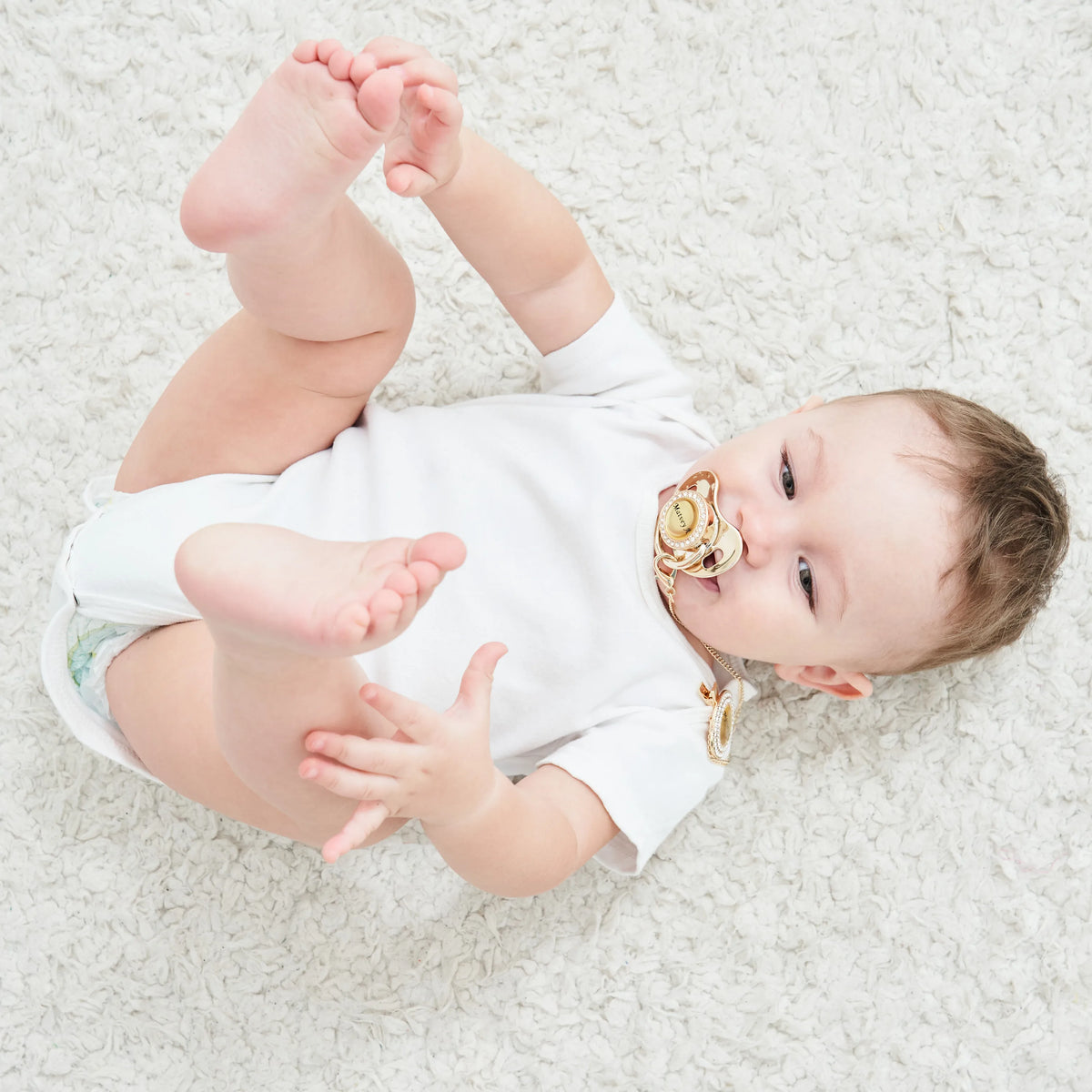 A baby lying on a white fluffy surface holding a personalized crystal pacifier and clip gift set