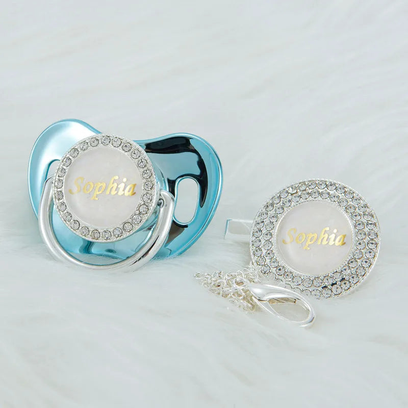A silver blue rhinestone pacifier clip set featuring a white disc with the name Sophia engraved on it