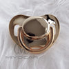 A gold-toned silicone pacifier with a smooth, rounded top, resting on white fluffy material.