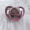 A silver and pink silicone pacifier with a rounded top is shown against a white, fluffy background.