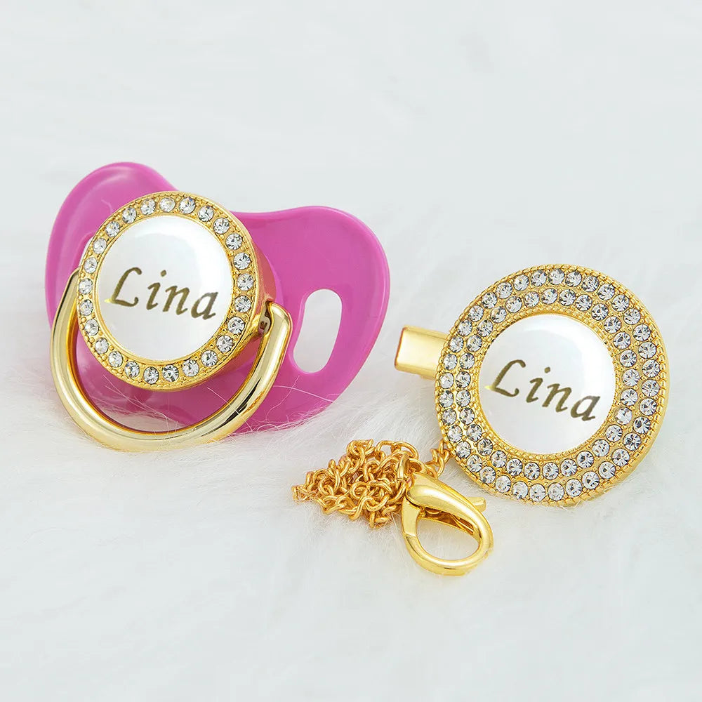 A pink pacifier clip with a gold ring and a white name charm featuring the name Lina and sparkling stones.