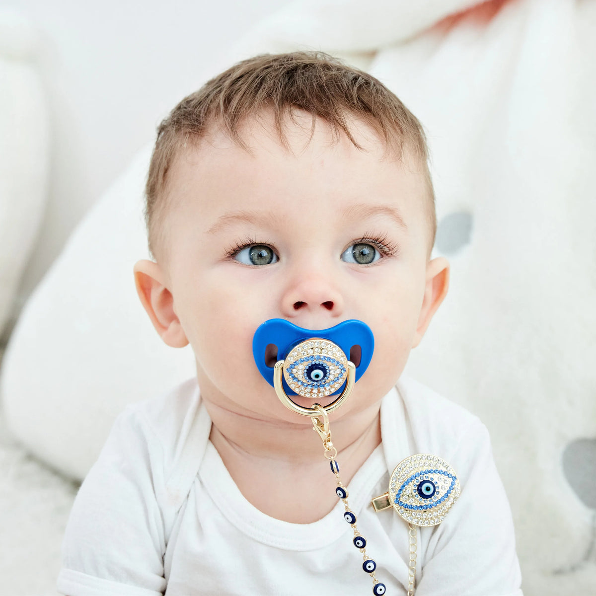 A baby is shown holding a blue evil eye pacifier and clip set.