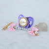 A purple crystal pacifier clip with a gold chain and pink ribbon, featuring the name Lina.