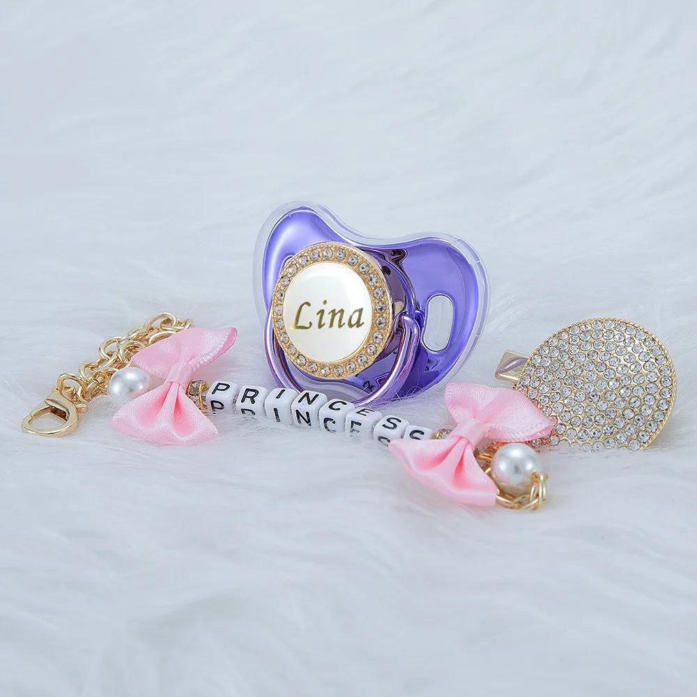 A purple crystal pacifier clip with a gold chain and pink ribbon, featuring the name Lina.