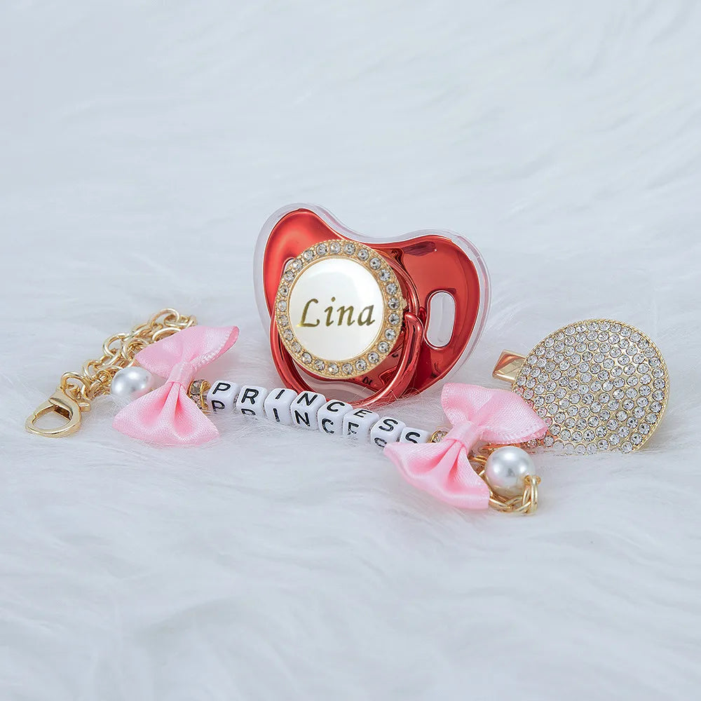 A personalized crystal pacifier with a pink clip and matching pearl charm resting on white fur.