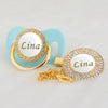 A gold pacifier clip with a light blue silicone holder and a circular white nameplate featuring the name Lina and gold bling.