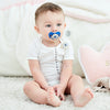 A baby is sitting on a white fluffy surface holding a blue evil eye pacifier and clip set.